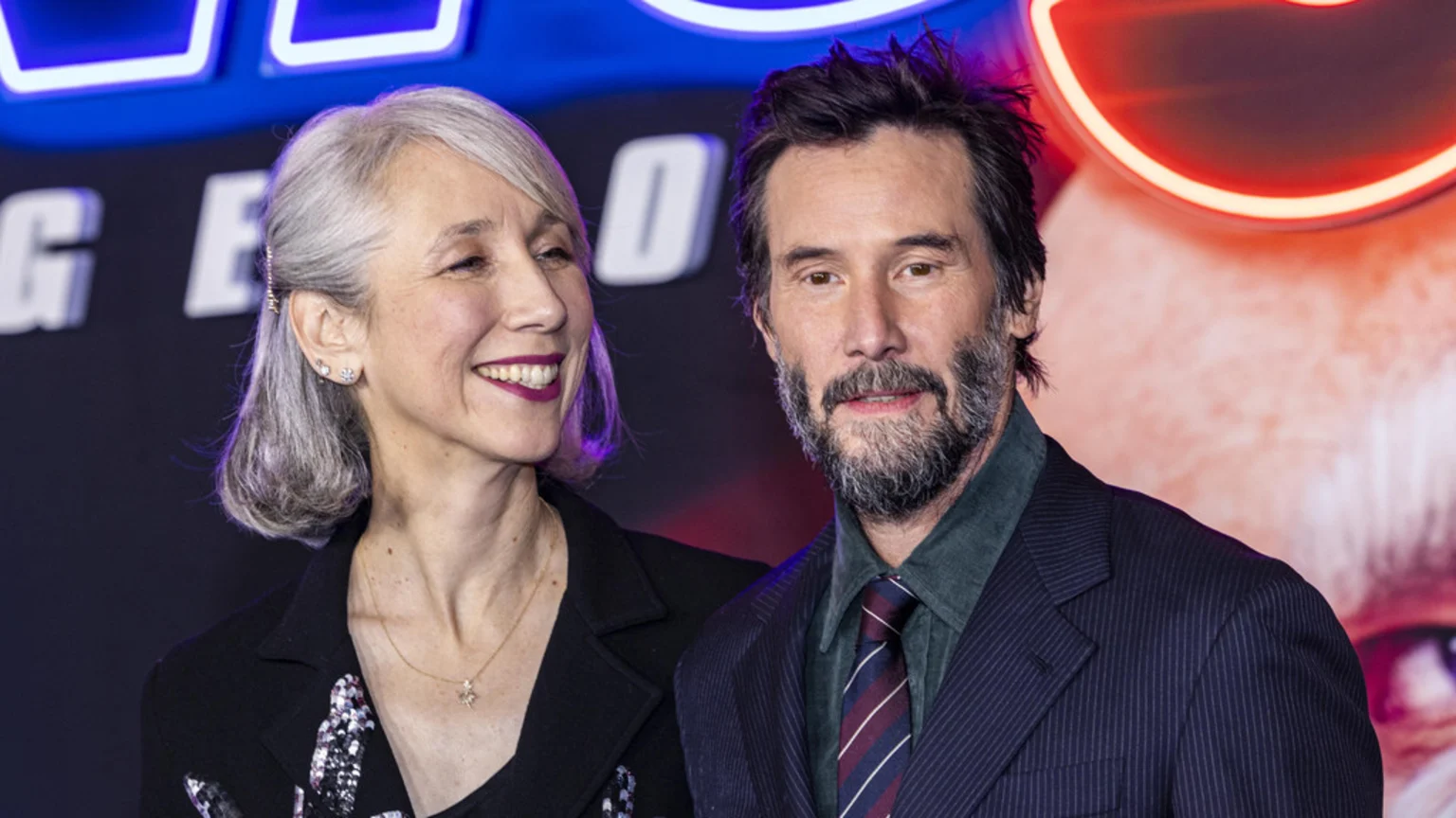 Keanu Reeves, with a beard and dark suit, looks at the camera while standing next to Alexandra Grant, who has grey hair and smiles towards him, with blurred neon signs in the background.