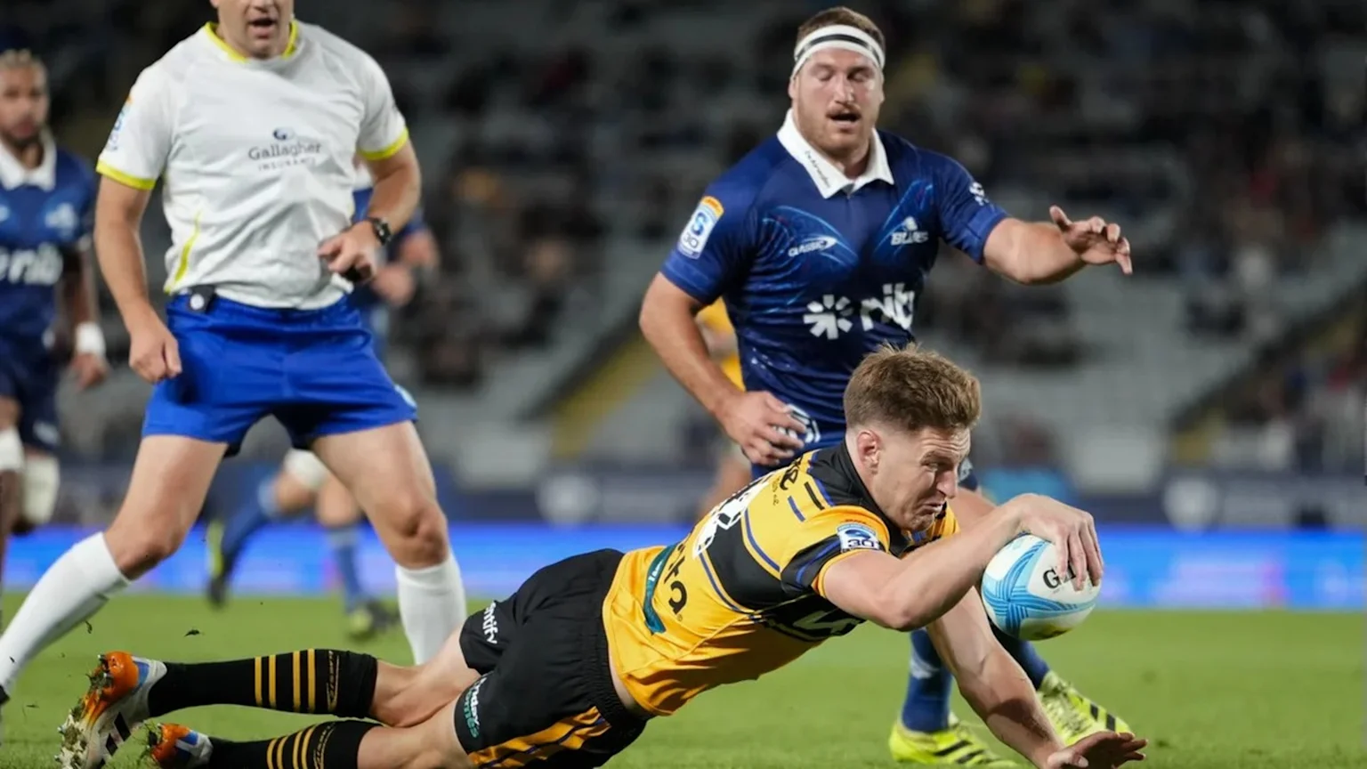 A rugby player in a yellow and black jersey dives to score a try with a white and blue ball, watched by a player in a blue jersey and a referee in a white shirt with a 'Gallagher' logo.