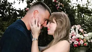 Taylor Swift holding Travis Kelce's cheeks with their foreheads pressed together surrounded by white and pink flowers in a garden