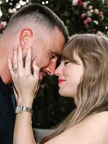 Taylor Swift holding Travis Kelce's cheeks with their foreheads pressed together surrounded by white and pink flowers in a garden