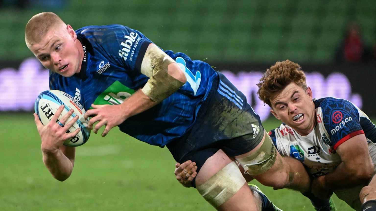 A rugby player in a blue jersey falls forward with the ball, being tackled by an opponent in a white, blue, and red jersey on a green field.