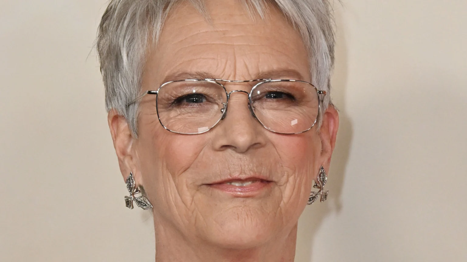 A close-up headshot of Jamie Lee Curtis smiling, with short gray hair, wearing silver-framed eyeglasses, and ornate silver leaf earrings.