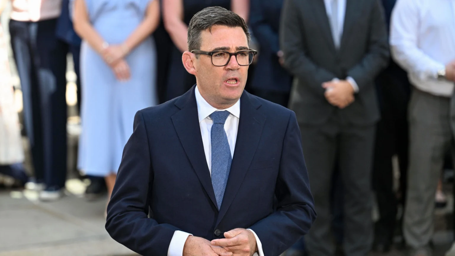A man in glasses, a dark blue suit, white shirt, and blue patterned tie speaks with his hands clasped in front, as blurred people stand behind him.