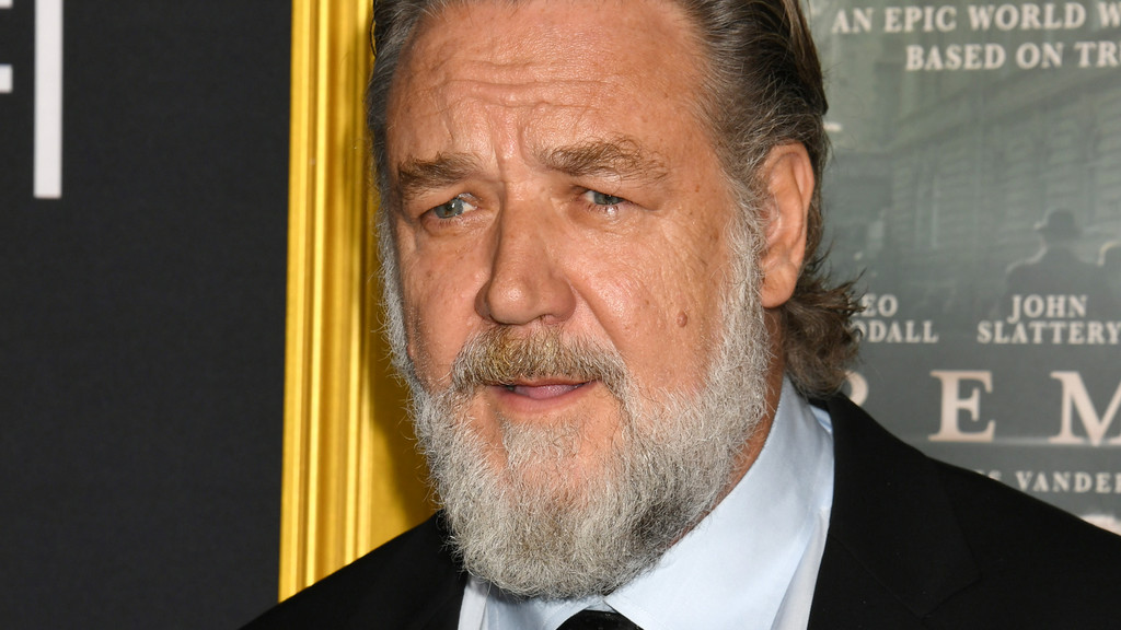 A close-up portrait of a man with a full grey beard and mustache, blue eyes, and grey hair, wearing a light blue shirt and dark suit jacket, with a slight smile, against a blurry background featuring a golden frame on the left and a poster on the right.