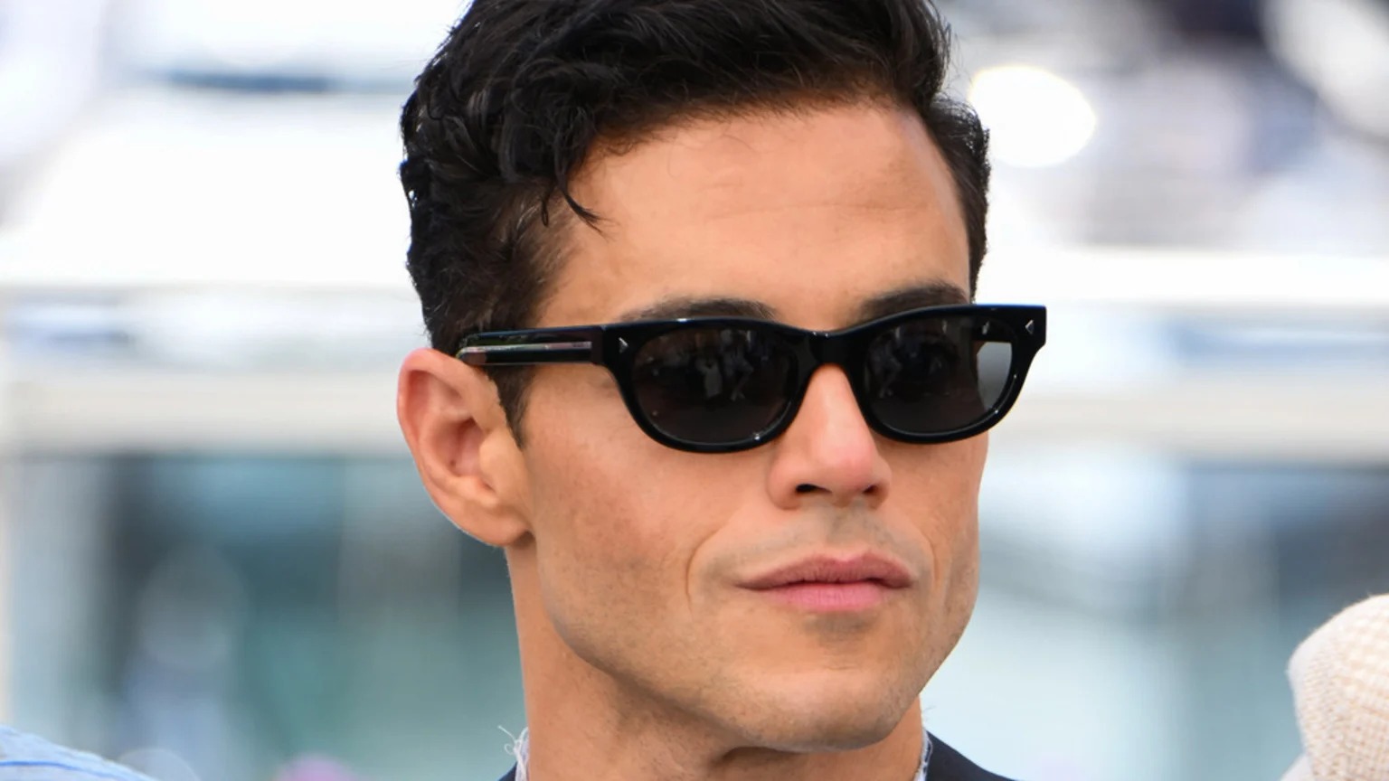 Close-up of actor Rami Malek wearing black sunglasses, with dark wavy hair, looking slightly to the right with a subtle expression.