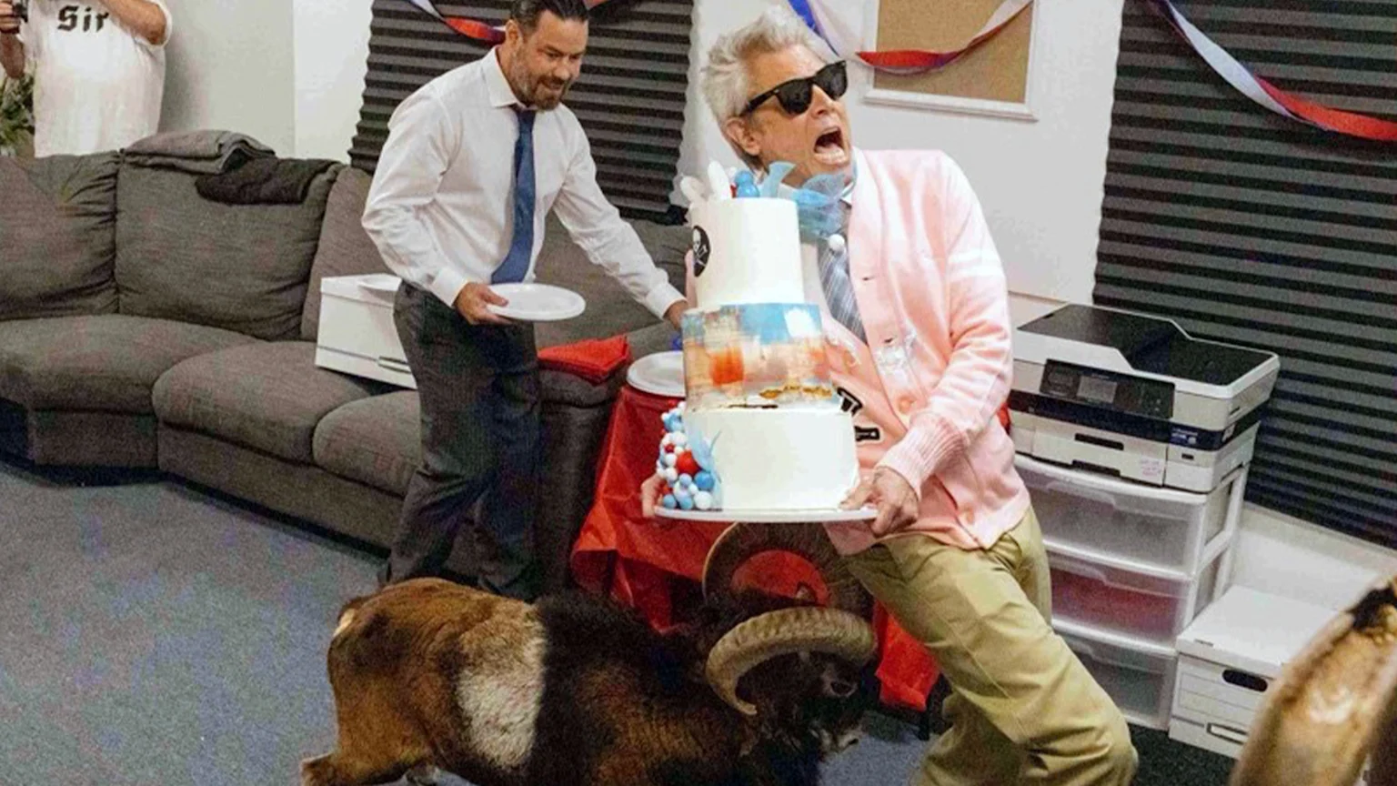Startled Johnny Knoxville in sunglasses and a pink cardigan holds a multi-tiered cake as a ram stands near his feet, while a man in a tie smiles in the background.