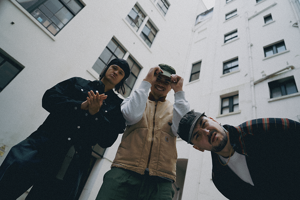 Three men pose outdoors from a low angle against a white building; one clasps his hands, another smiles while adjusting his hat, and the third leans into the frame.