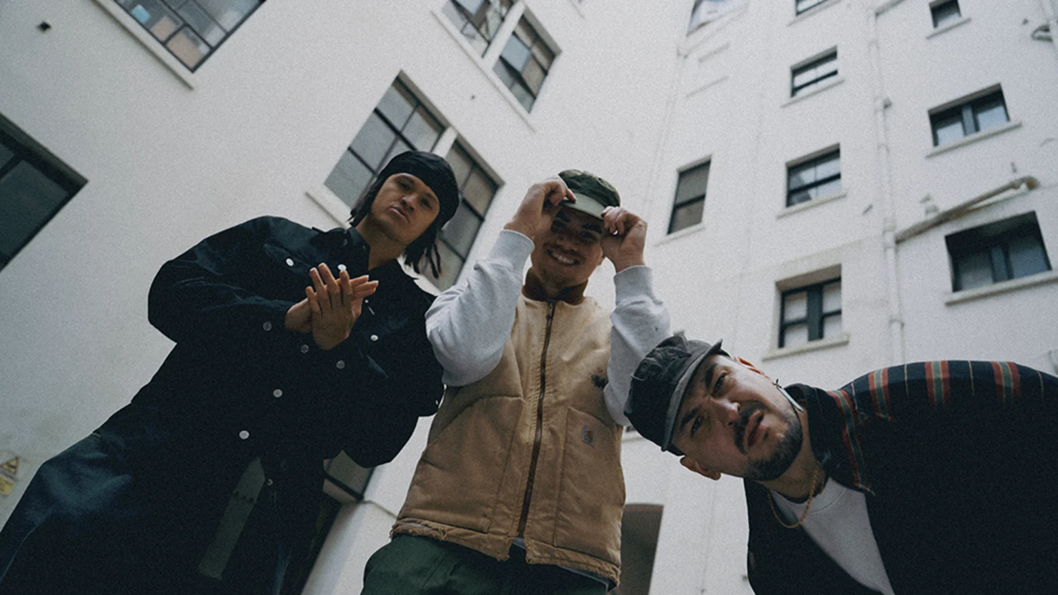 Three men pose outdoors from a low angle against a white building; one clasps his hands, another smiles while adjusting his hat, and the third leans into the frame.