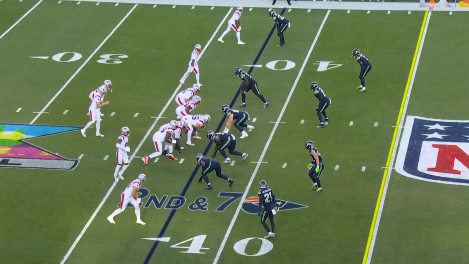 Overhead view of an American football game on a green turf field with players in white/red and dark green/lime green uniforms. Visible are 30 and 40 yard lines, a '2ND & 7' down marker, New England Patriots and NFL logos, yellow first down and blue line of scrimmage lines, plus a colorful abstract logo.
