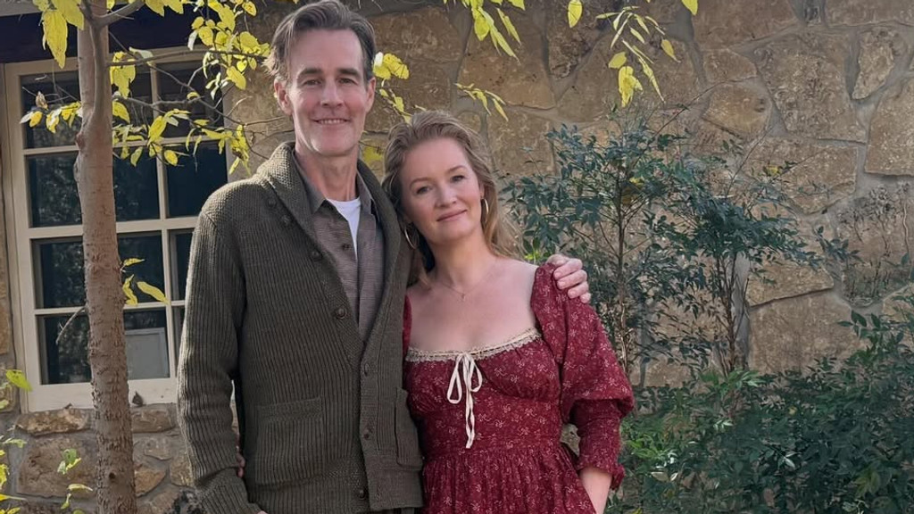 A smiling man in an olive green cardigan with his arm around a woman in a red floral dress with puff sleeves and lace trim. They stand outdoors in front of a stone wall and green foliage, with a tree displaying yellow leaves and a window visible on the left.