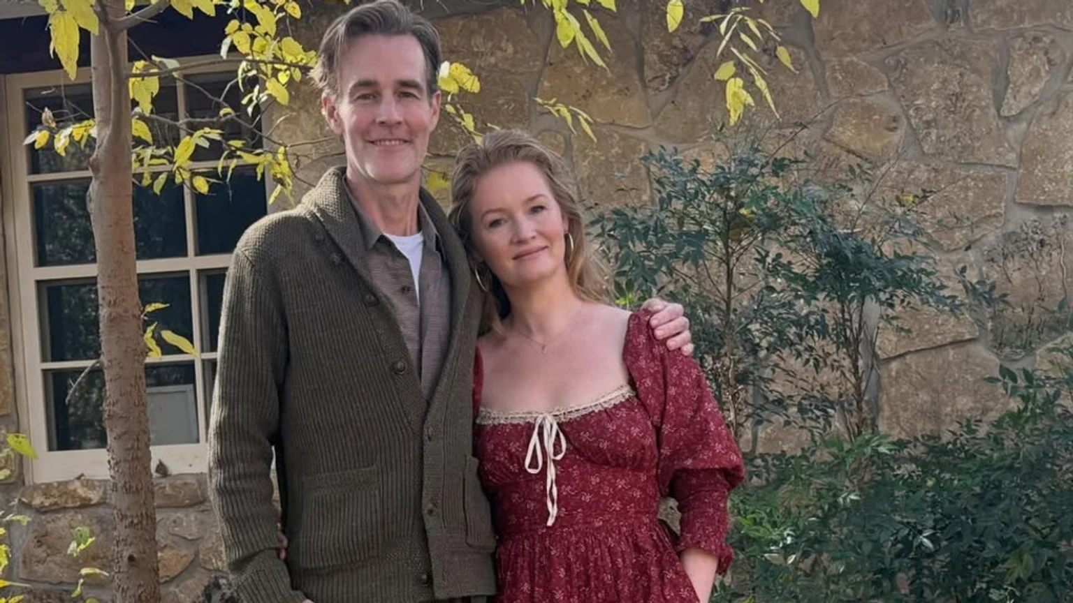 A smiling man in an olive green cardigan with his arm around a woman in a red floral dress with puff sleeves and lace trim. They stand outdoors in front of a stone wall and green foliage, with a tree displaying yellow leaves and a window visible on the left.