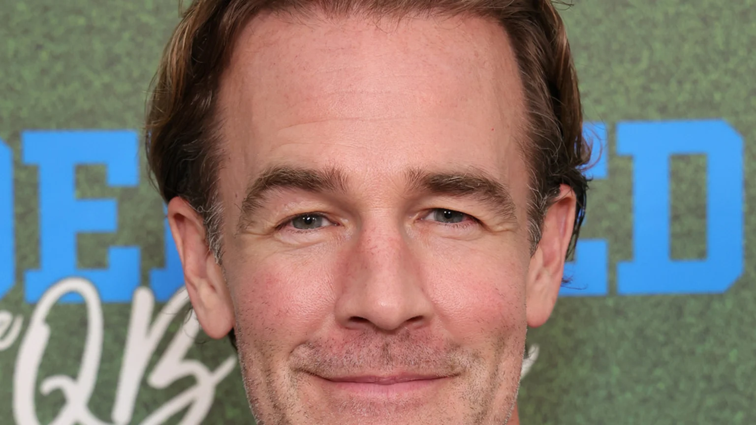 A close-up portrait shows actor James Van Der Beek, a light-skinned man with brown hair and blue eyes, smiling faintly at the camera against a blurry green and blue background.