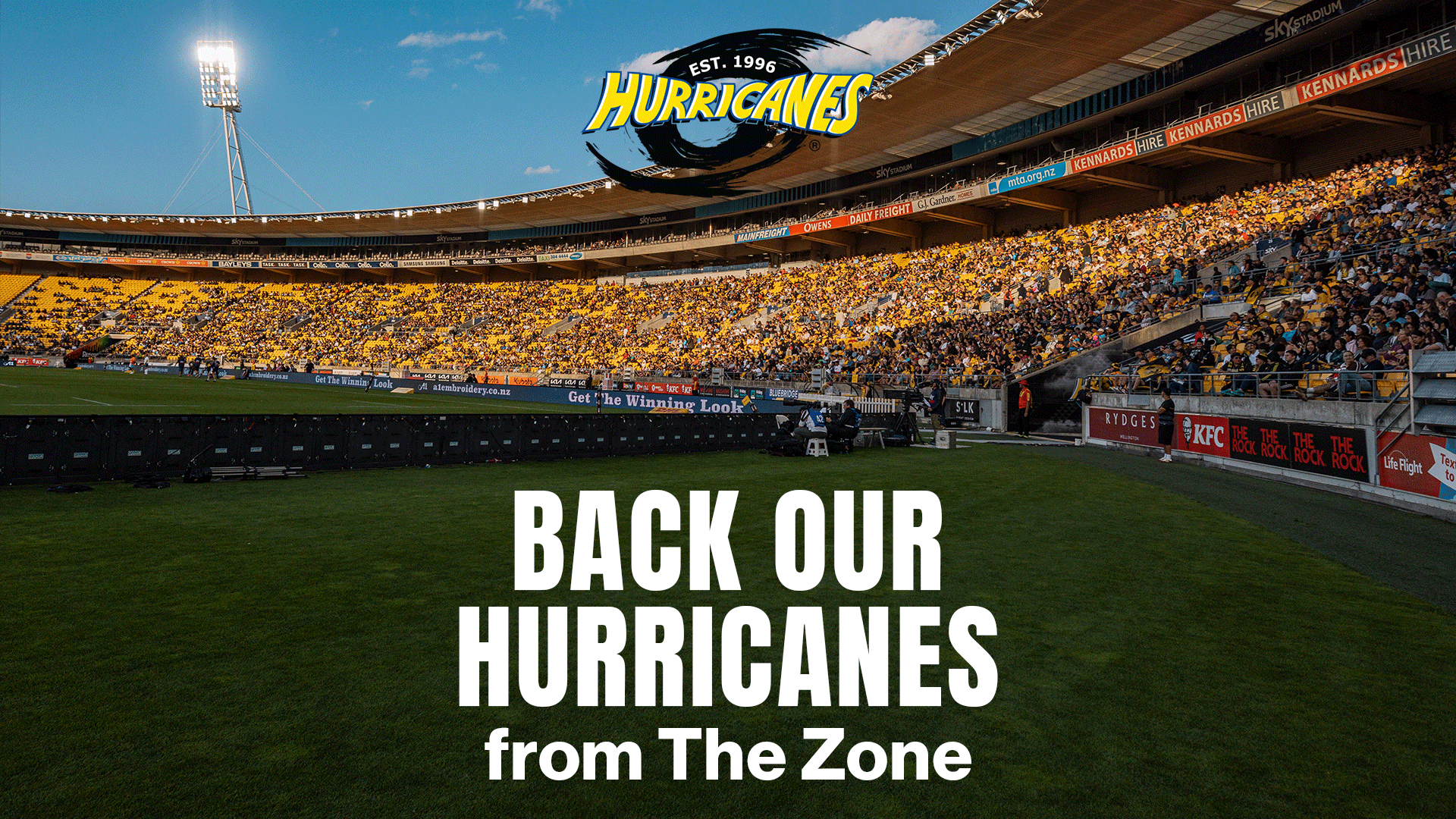 A large stadium is packed with fans wearing yellow and black, viewed from the field level, with a prominent Hurricanes logo and "BACK OUR HURRICANES from The Zone" text.