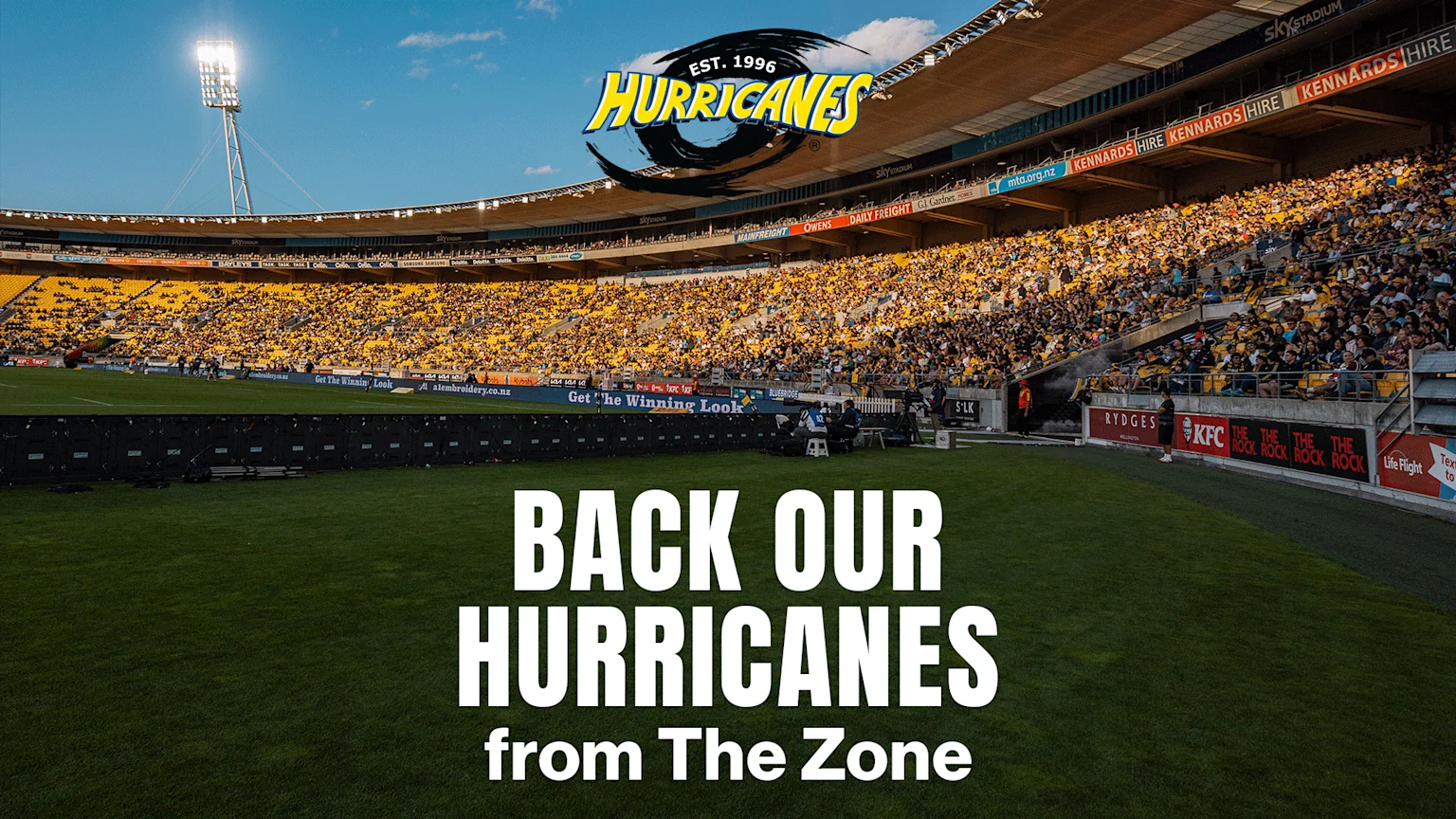 A large stadium is packed with fans wearing yellow and black, viewed from the field level, with a prominent Hurricanes logo and "BACK OUR HURRICANES from The Zone" text.