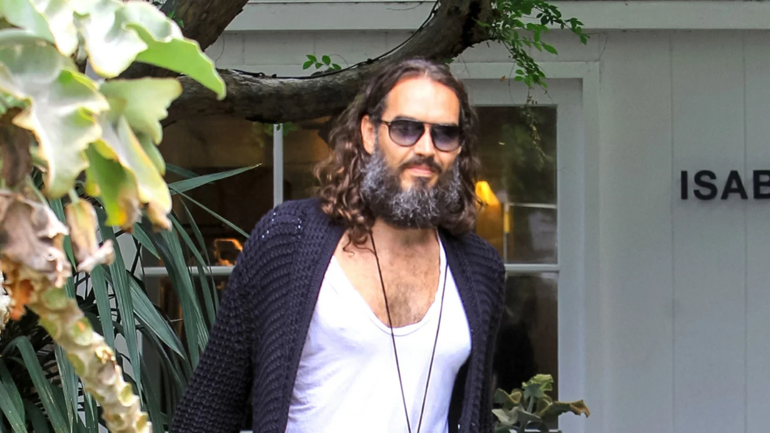 Russell Brand, with long curly hair, a grey beard, and sunglasses, wears an open dark cardigan over a white tank top, standing in front of a white building with plants.