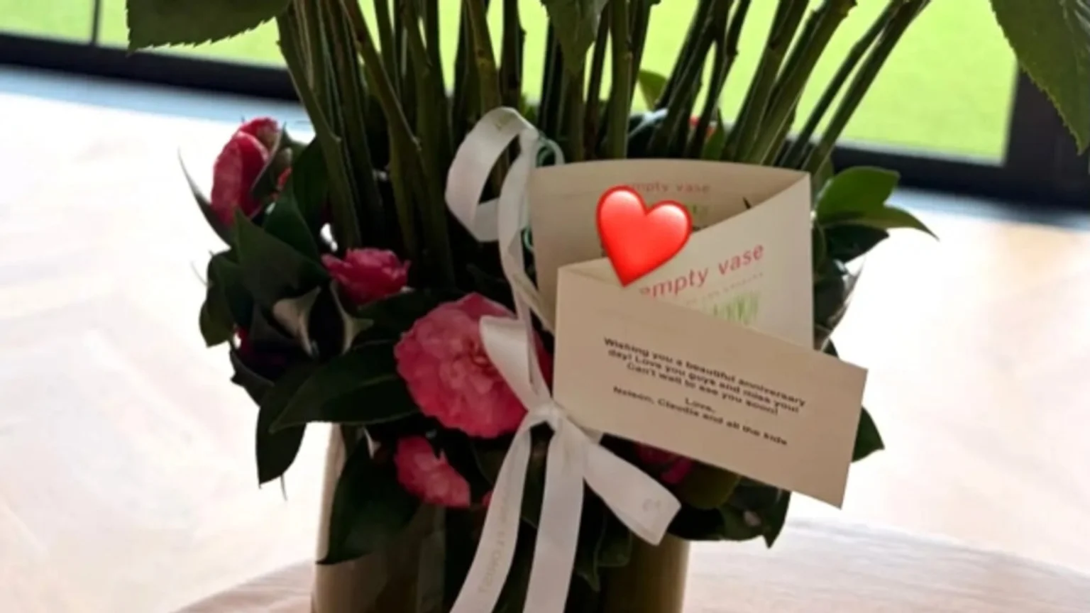 A bouquet of pink flowers and green foliage in a glass vase, adorned with white ribbons and two attached cards; one card has a red heart emoji, and the other shows a handwritten anniversary message.