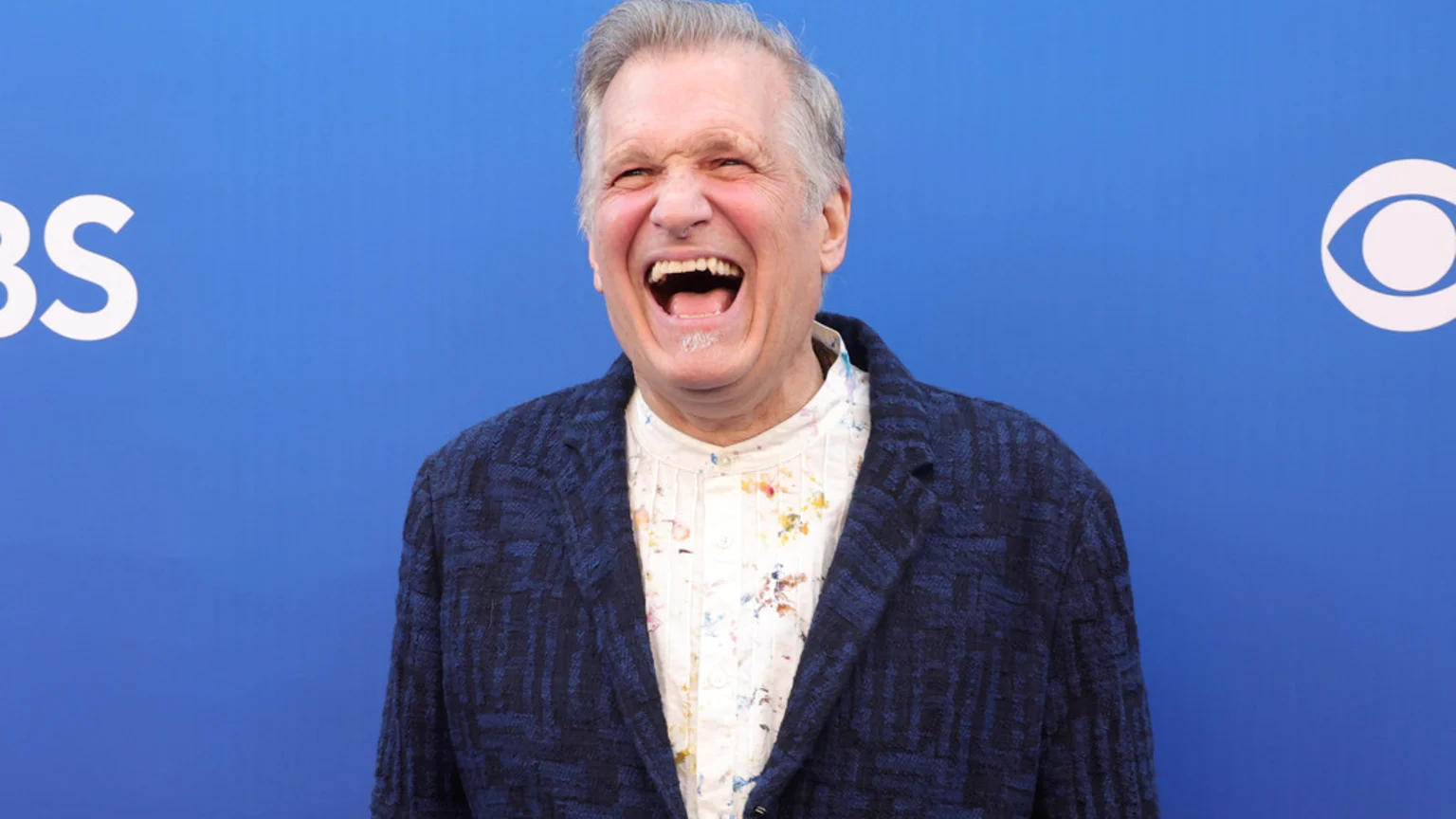 Drew Carey laughs loudly, wearing a blue patterned jacket over a white, paint-splattered shirt, against a blue background featuring CBS logos.