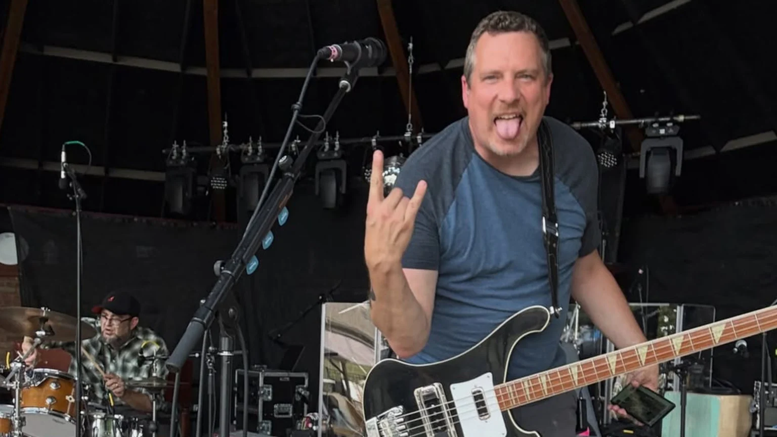 A male bassist in a blue shirt sticks his tongue out and makes a rock-on gesture while playing a black bass guitar on stage, with a drummer visible in the background.
