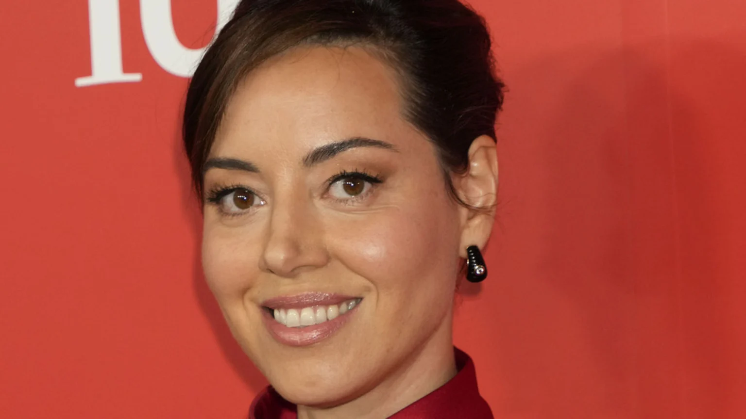 A close-up of Aubrey Plaza smiling, wearing a red top and a black earring against a red background with a partial white letter in the upper left.