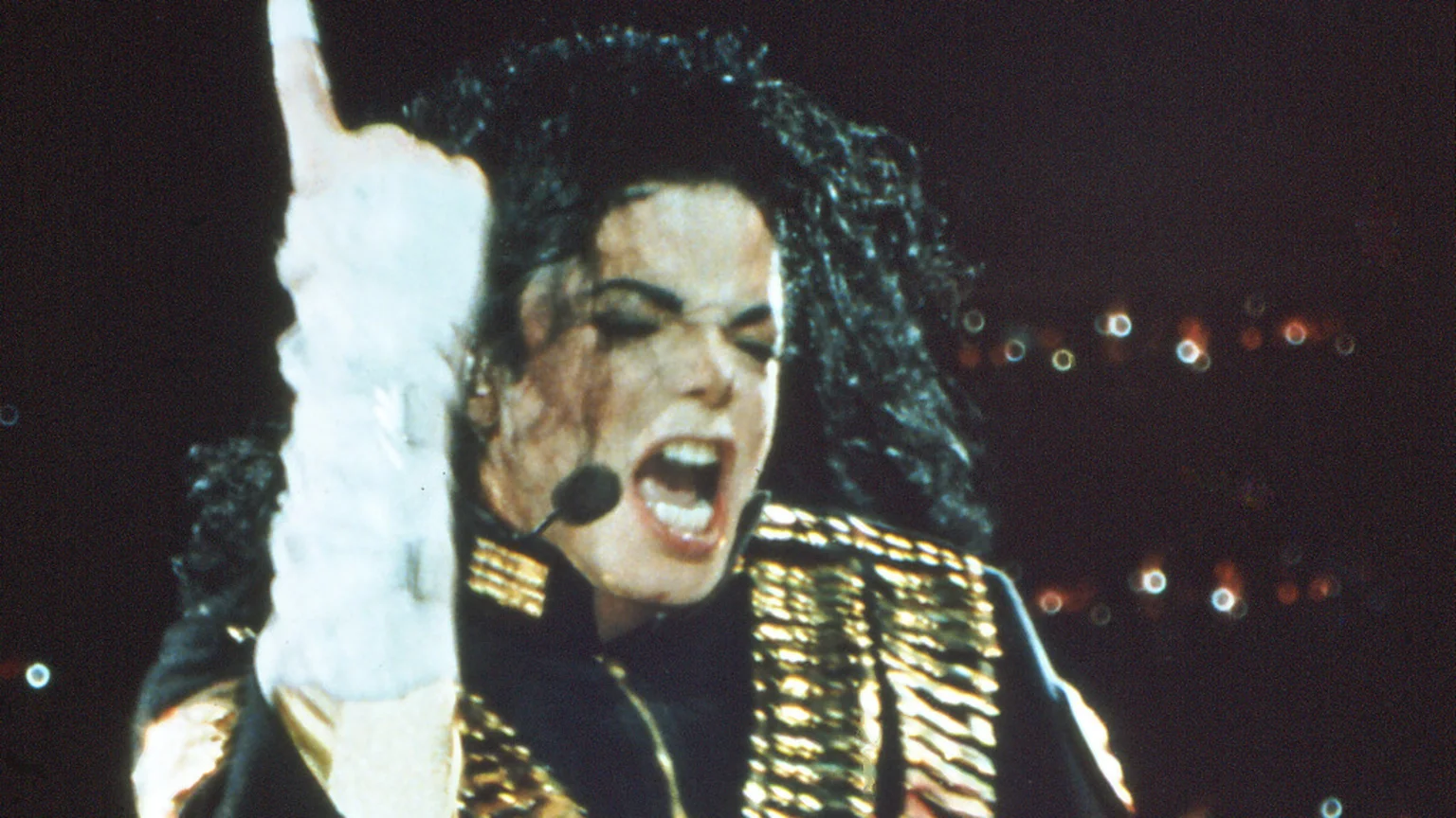 Michael Jackson on stage, wearing a black and gold jacket, white glove, and headset microphone, with his mouth open in song and left hand pointing up, against a dark background with blurred lights.