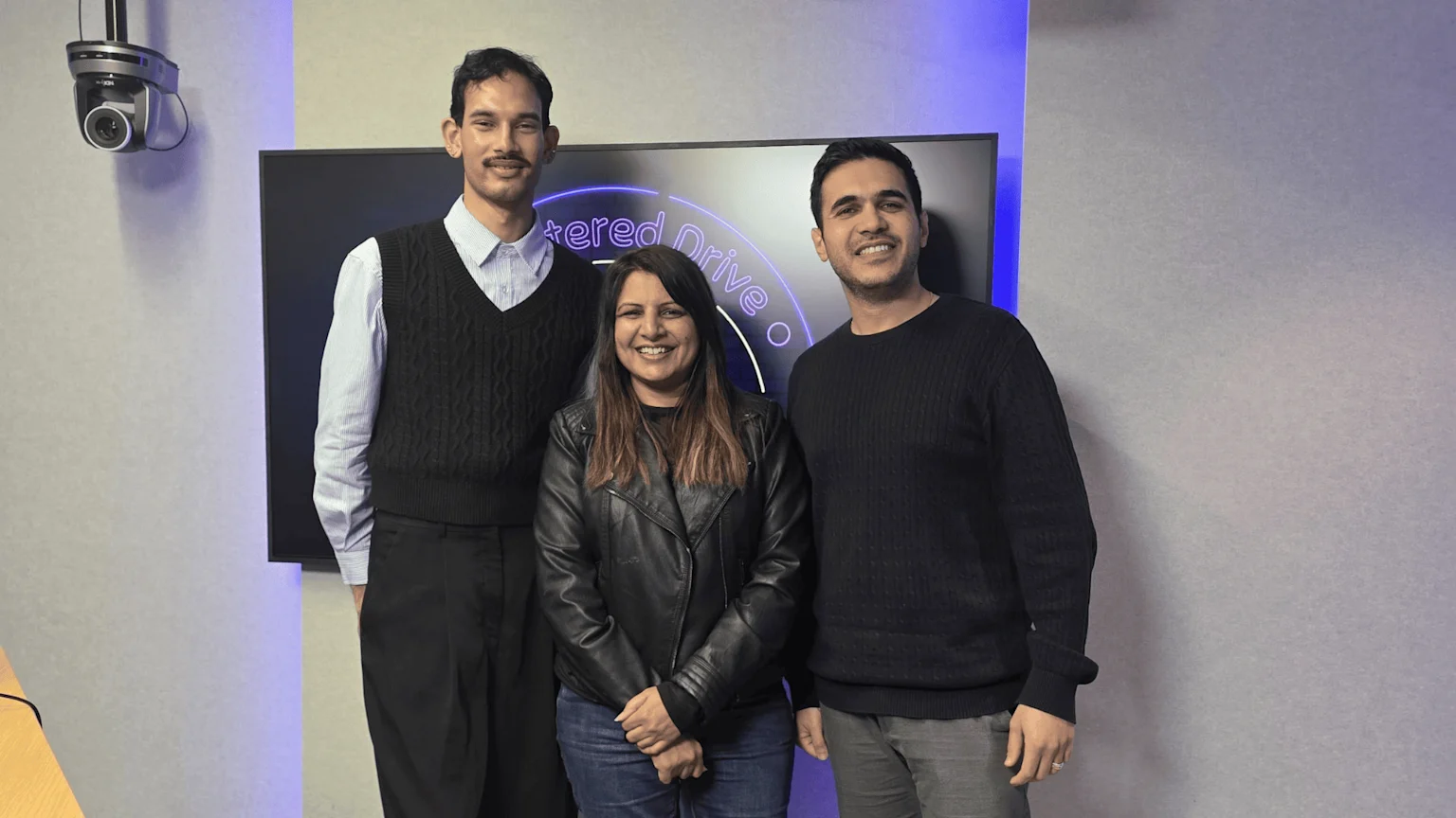 Three smiling people, two men and one woman, stand in a studio in front of a screen with partial text "tered Drive o", with a camera mounted on the wall to the left.