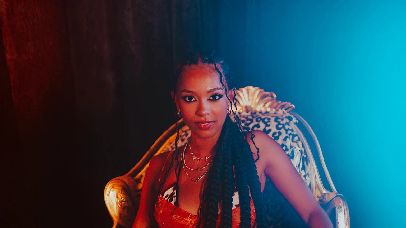 A young woman with braided hair and dark skin sits in an ornate, leopard-print chair, looking at the camera, illuminated by red and blue light.