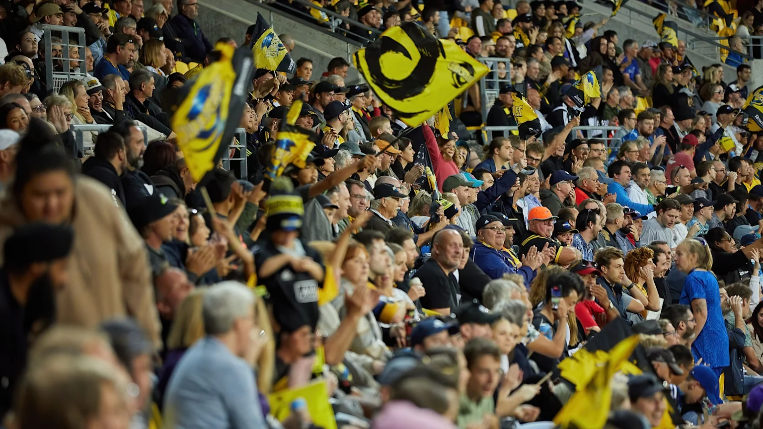 A large, animated crowd fills a stadium, with many spectators wearing yellow and black attire and waving yellow flags with various logos.