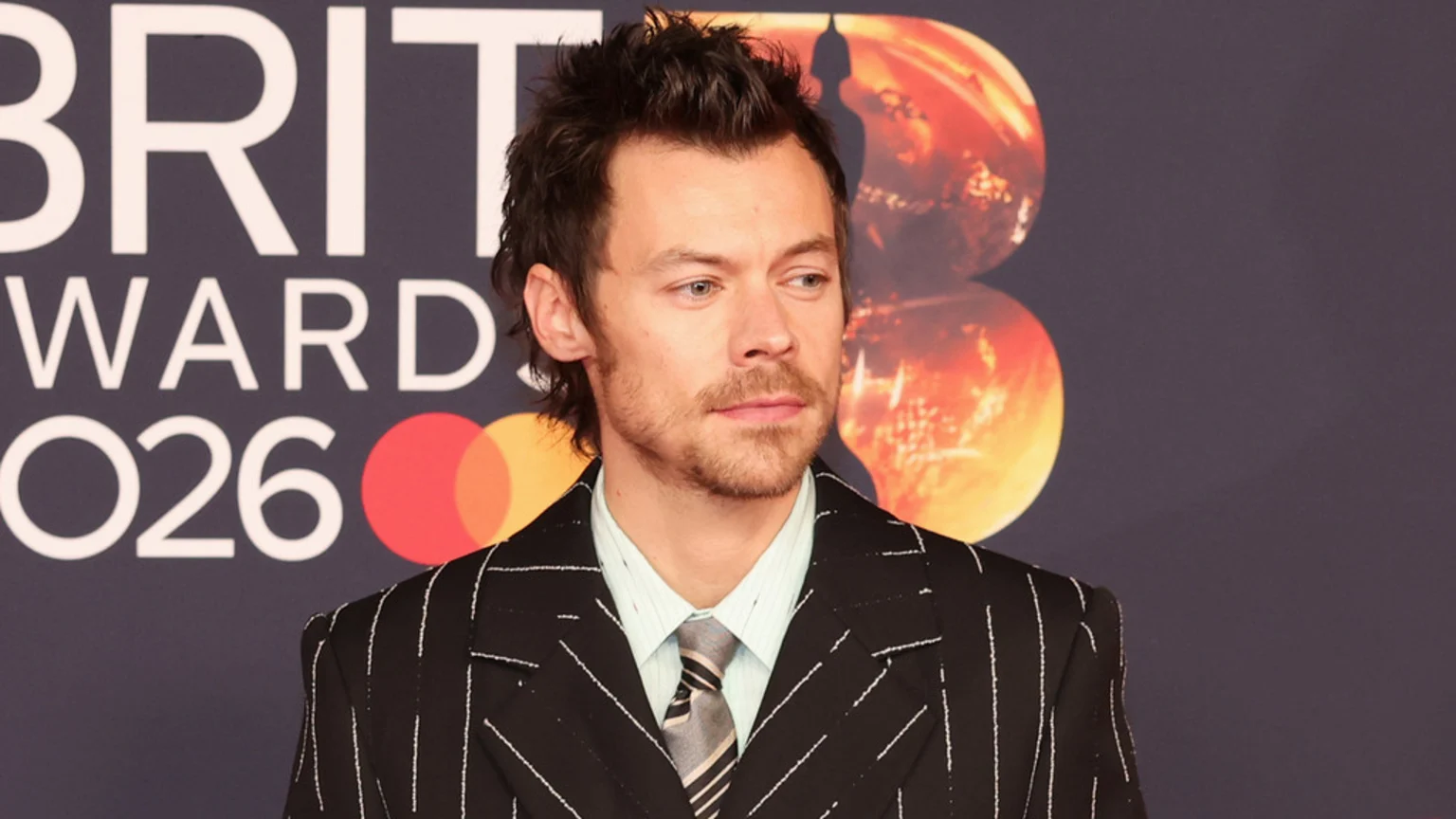 Harry Styles, with dark messy hair and stubble, wears a black pinstripe suit jacket, light blue striped shirt, and striped tie, posing against a "BRIT AWARDS 2026" backdrop.