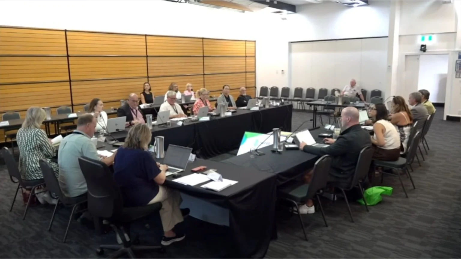 A group of approximately 20 people are seated around U-shaped tables in a conference room, many looking at laptops, while one man stands at the far end, addressing the attendees.