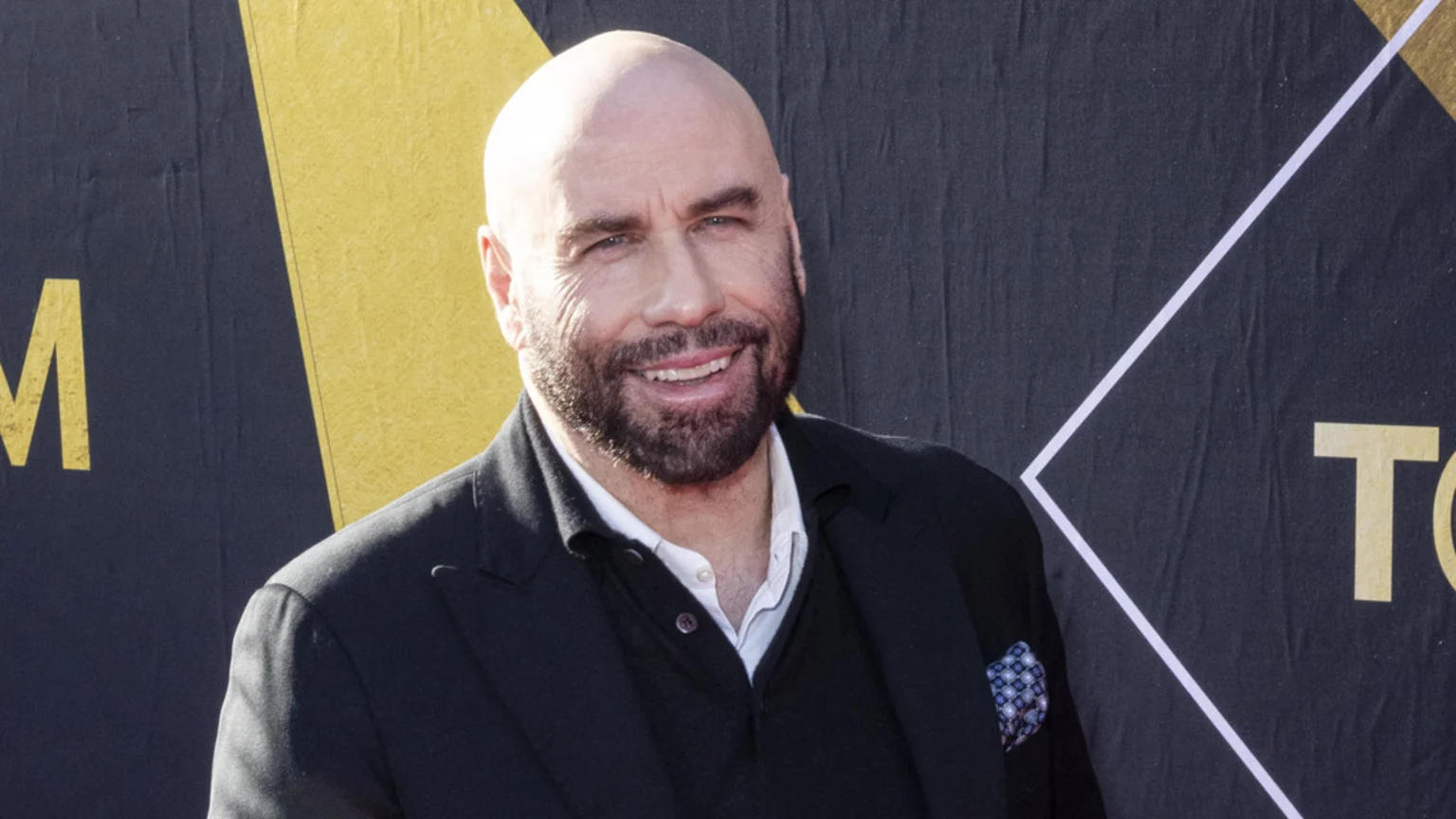 John Travolta, bald with a beard, smiles broadly while wearing a dark suit jacket, dark collared shirt, and a pocket square, standing against a black and gold backdrop.