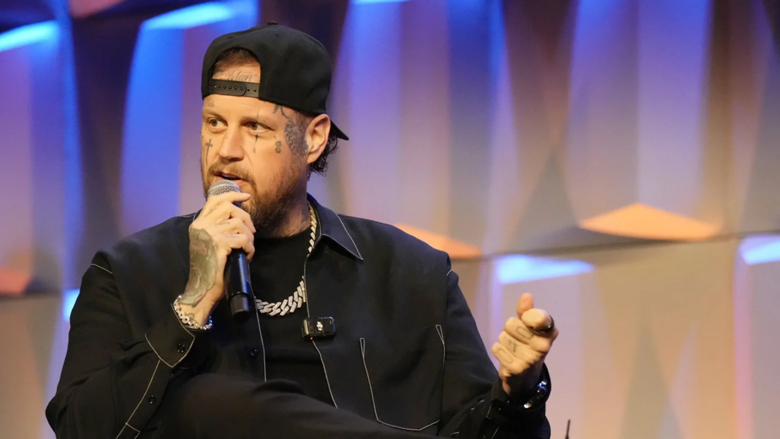 A bearded man with face and arm tattoos, wearing a backward black cap, black collared shirt with white stitching, thick silver chain, and bracelet, speaks into a microphone, with a lavalier mic clipped to his shirt, against a blurred background of blue and orange lights.