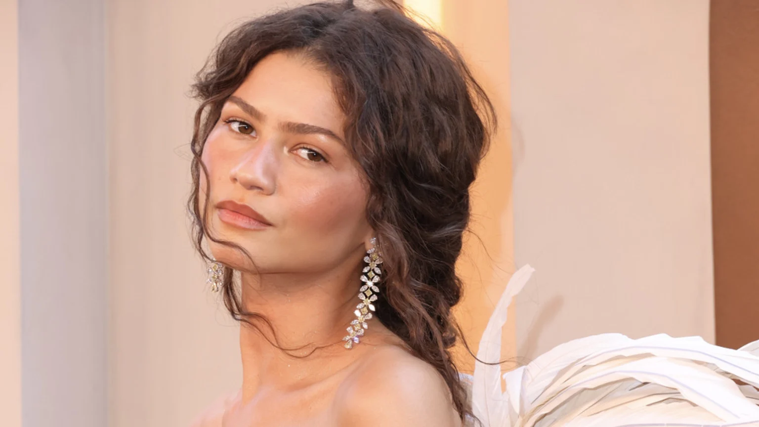 Close-up of Zendaya looking at the camera, wearing long, sparkling floral earrings and a white, layered outfit, with her dark, curly hair down.