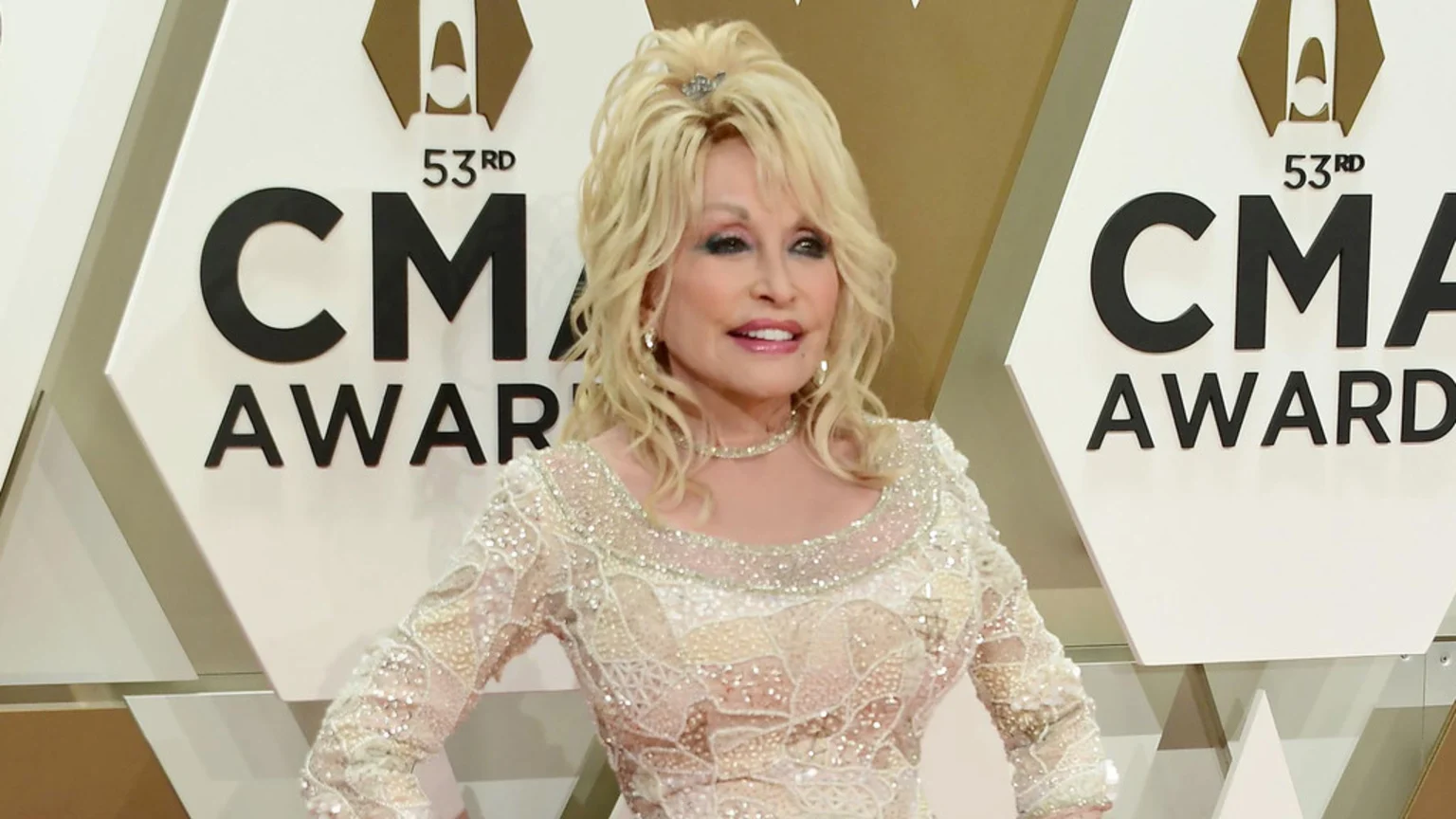Dolly Parton smiles, wearing a sparkly cream gown with a matching choker and earrings, and her blonde hair styled up, against a white background with the "53rd CMA AWARDS" logo.