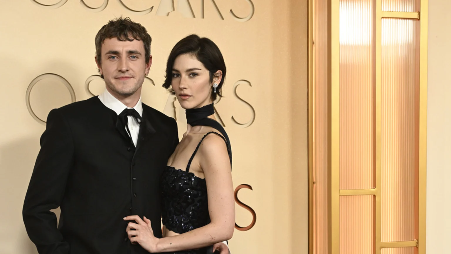 Paul Mescal in a black suit and Gracie Abrams in a black sequined top pose together against a beige wall with "OSCARS" visible in the background.