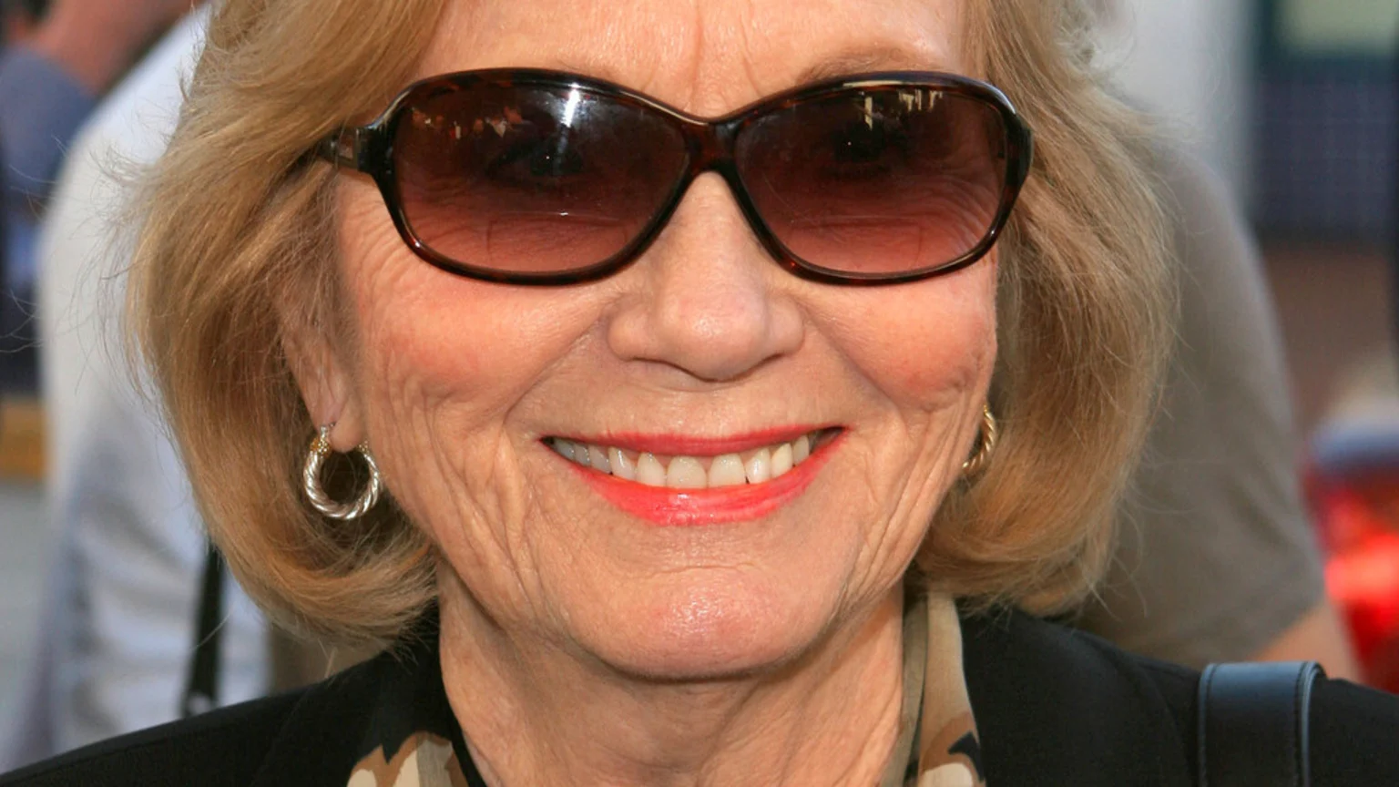 An older woman with blonde hair, dark sunglasses, and bright coral lipstick smiles, showing her teeth and silver hoop earrings.