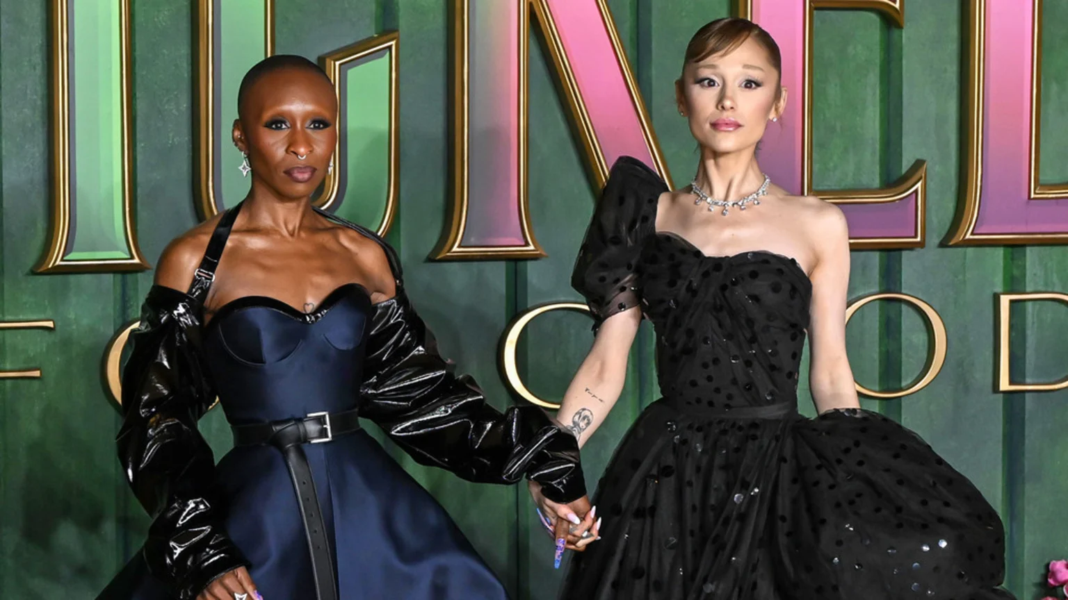Cynthia Erivo and Ariana Grande hold hands on a green carpet, Erivo in a dark blue corset dress with a black jacket, and Grande in a black strapless polka-dot dress.