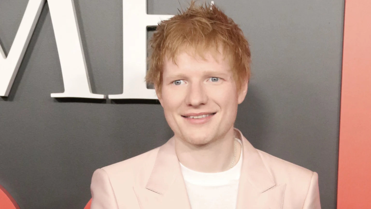 Smiling Ed Sheeran, with red hair and blue eyes, wears a light pink blazer over a white t-shirt, against a grey background with large white letters.
