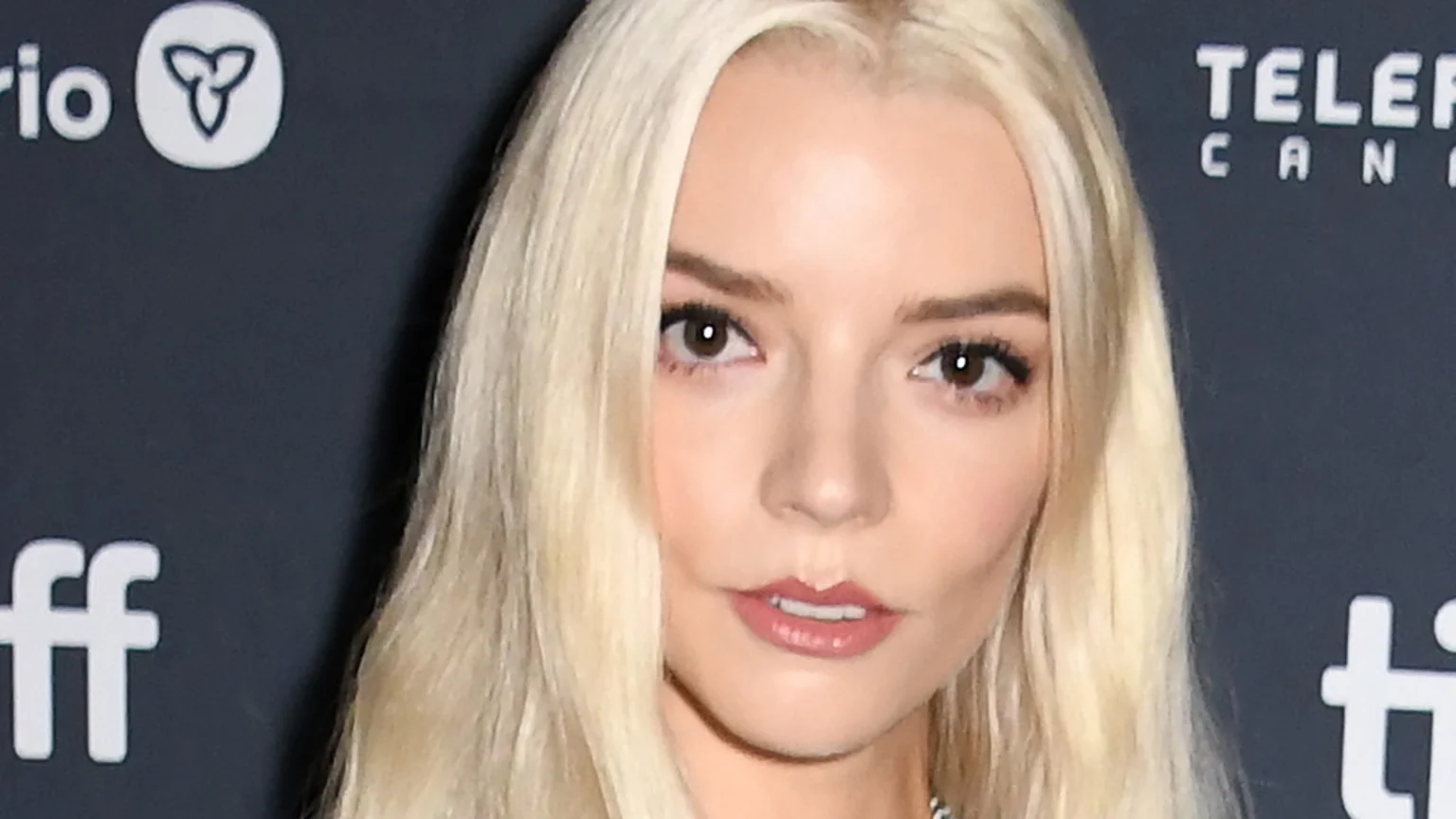 Close-up of Anya Taylor-Joy with long blonde hair and dark eyes, gazing directly at the camera. Blurred logos for 'TIFF' and 'Telefilm Canada' appear in the dark background.