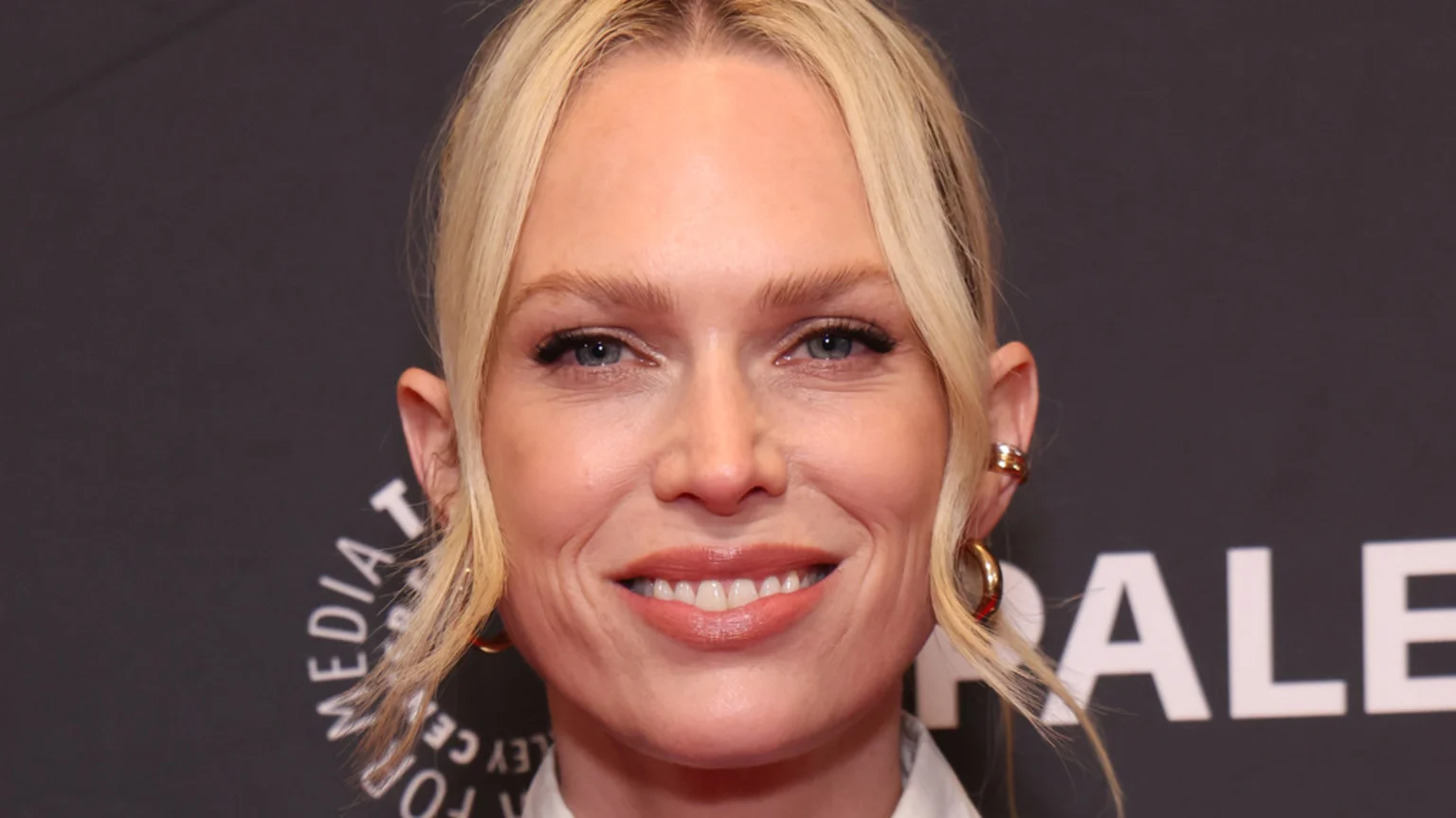 A smiling blonde woman with blue eyes and light makeup looks directly at the camera, wearing gold hoop earrings and a white shirt against a dark grey background with a partial white logo.