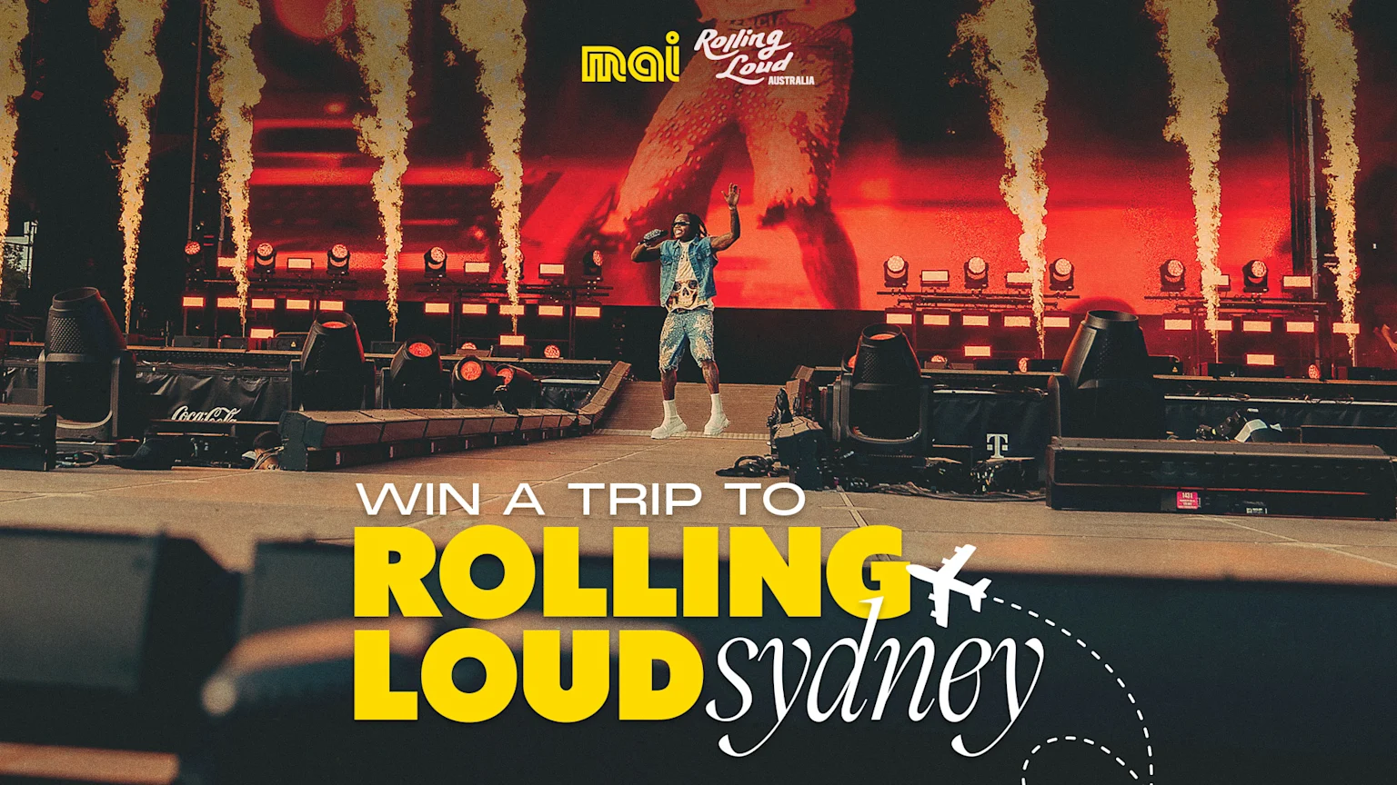 A vibrant concert scene from Rolling Loud Sydney featuring a male performer in blue denim and graphic t-shirt enthusiastically performing on stage with raised arms. The stage is dramatically lit with pyrotechnic flames, red backlighting, and a large LED screen displaying red and orange abstract imagery. Branding includes Coca-Cola, Rolling Loud Australia, and 'mai' logos. Overlay text invites viewers to win a trip to Sydney, accompanied by a white airplane icon with a dotted flight path.