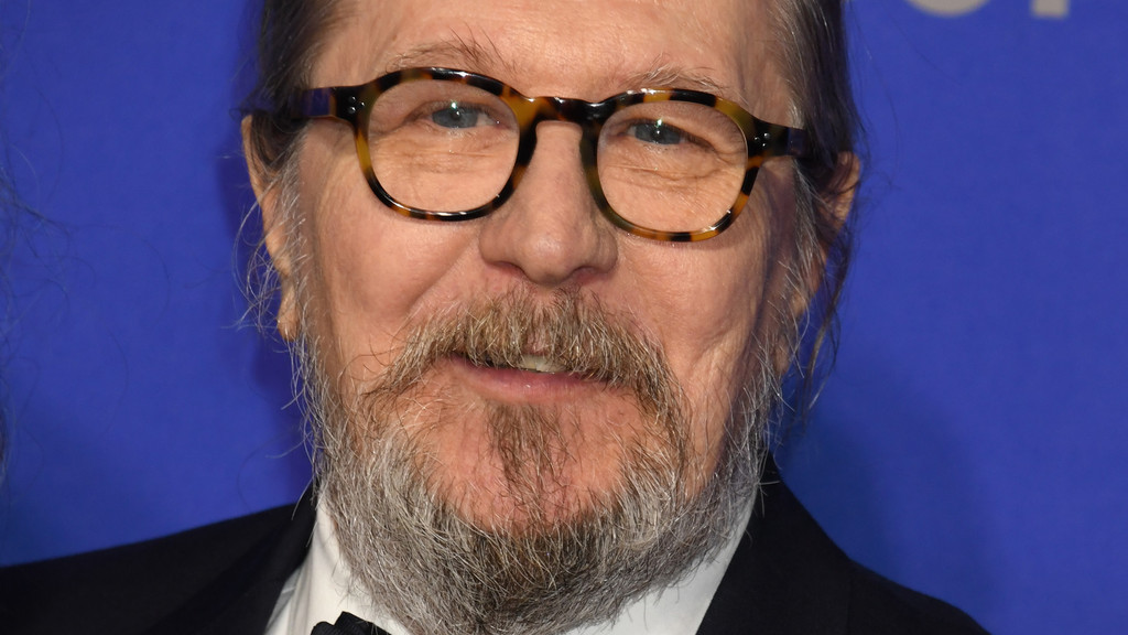 Close-up of actor Gary Oldman smiling, wearing tortoiseshell glasses, a gray beard, and a suit against a blue background.