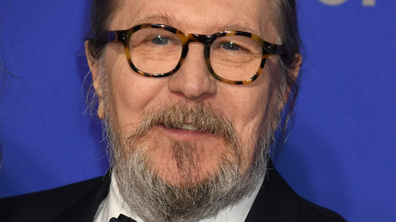 Close-up of actor Gary Oldman smiling, wearing tortoiseshell glasses, a gray beard, and a suit against a blue background.