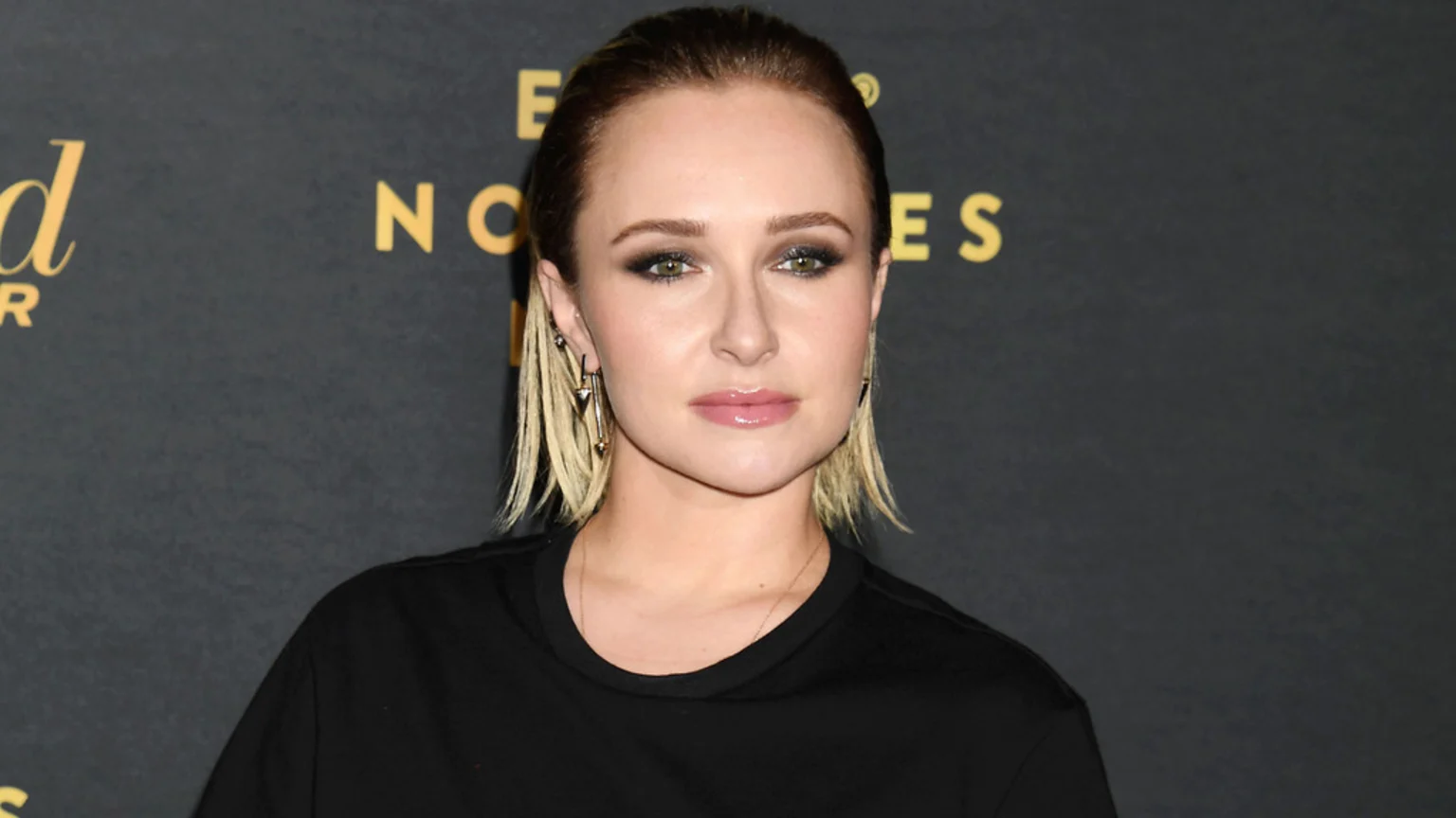 A woman with pulled-back dark hair, blonde streaks, dark eye makeup, and light pink lips looks directly at the camera, wearing a black top and geometric earrings against a dark background with gold lettering.