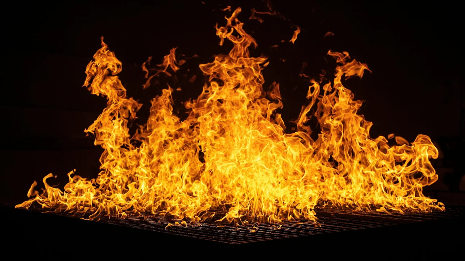 Intense orange and yellow flames spread across a dark metal grill against a black background.