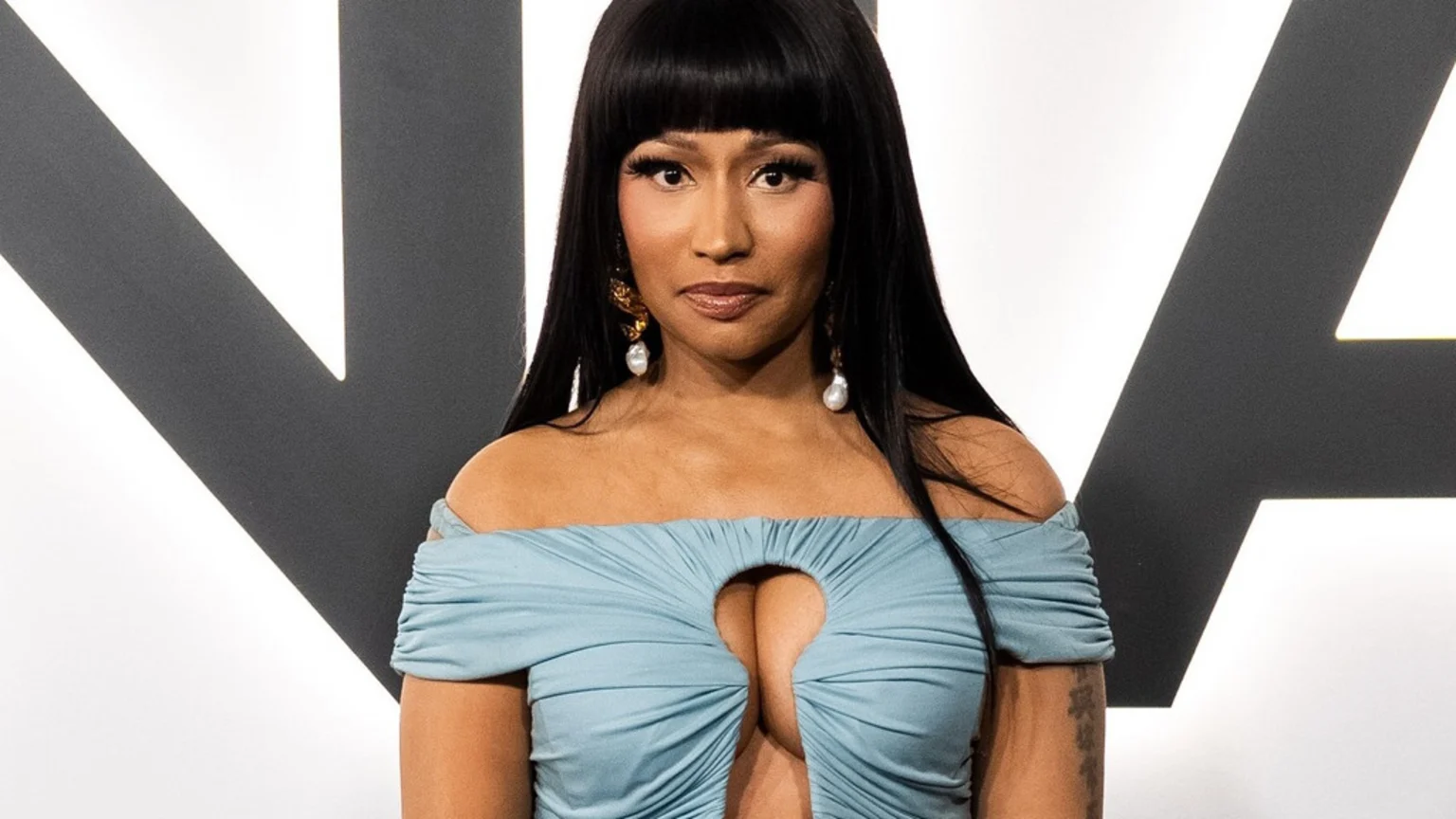 Nicki Minaj with long dark hair and bangs, wearing a light blue off-the-shoulder top with a chest cutout, and large pearl earrings, against a white background with dark geometric shapes.