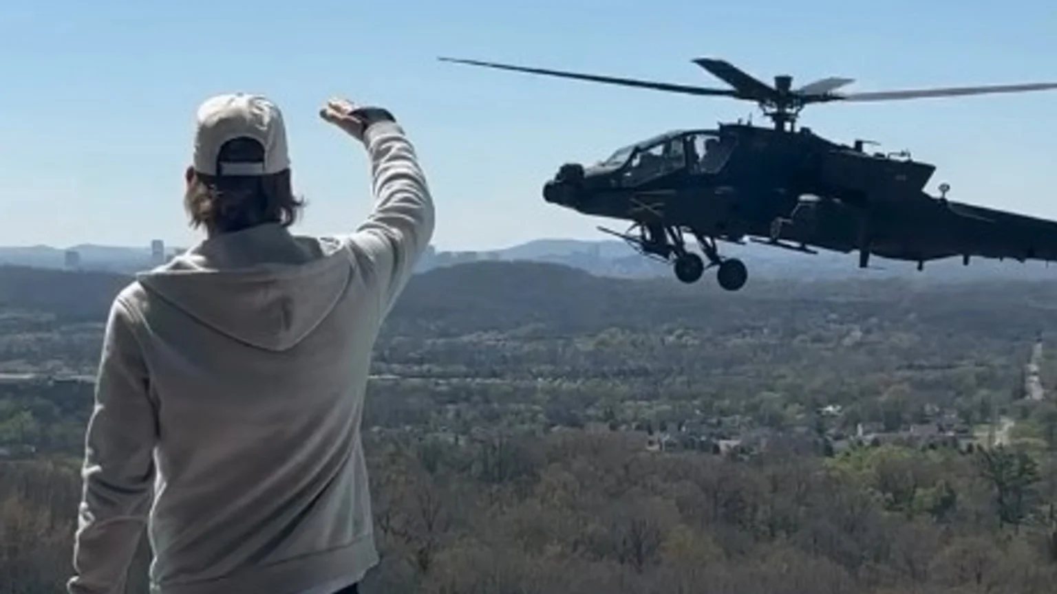 From behind, a person in a baseball cap and hoodie waves at a dark attack helicopter flying over a vast, tree-covered landscape with distant city buildings under a blue sky.