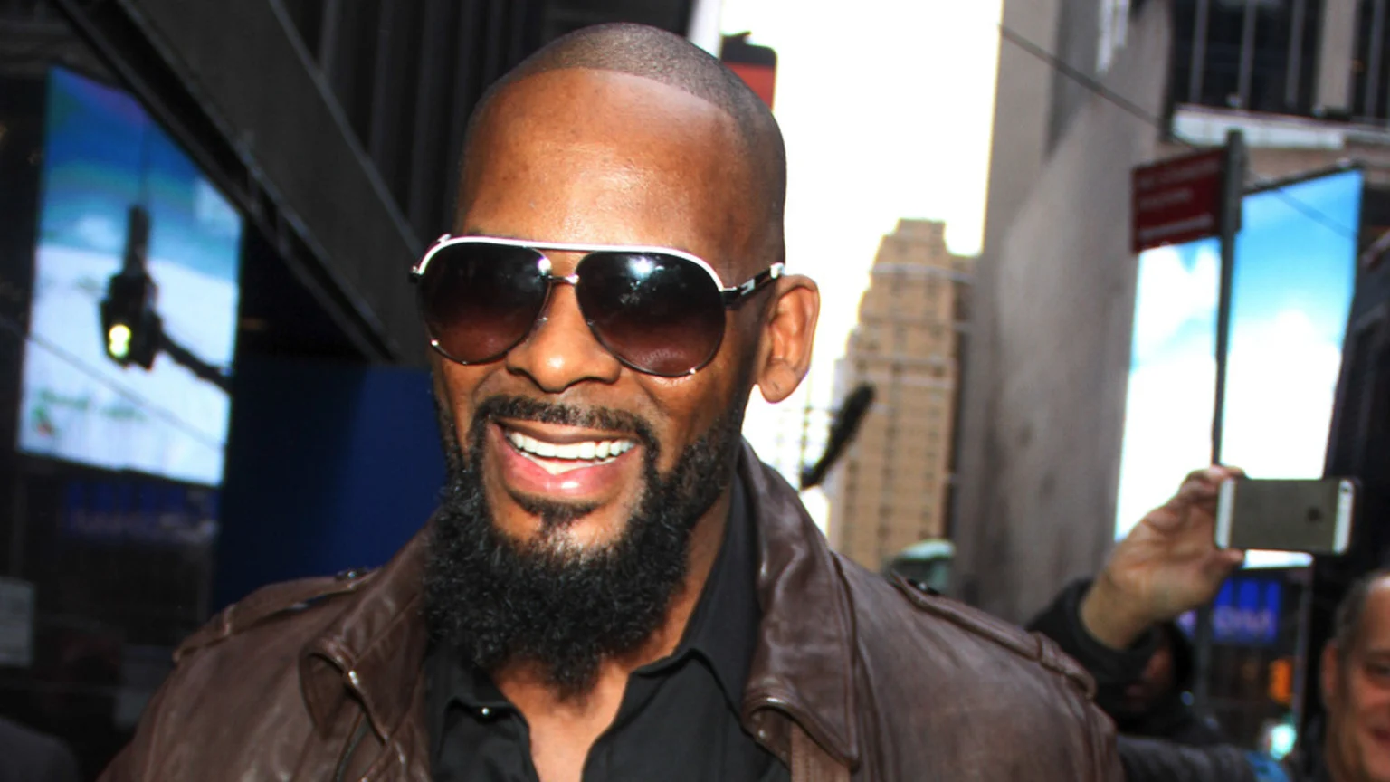 R. Kelly, a bald man with a full beard and aviator sunglasses, smiles broadly while wearing a brown leather jacket in a city street.