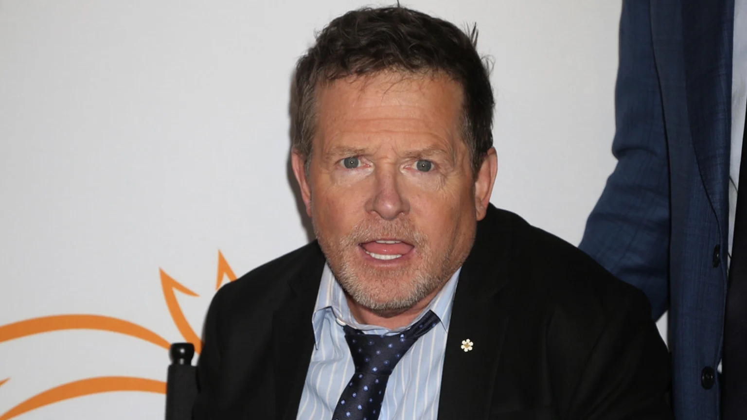 Michael J. Fox, with short hair and a beard, looks at the camera while wearing a black suit, striped shirt, and patterned tie, with a white flower pin on his lapel.