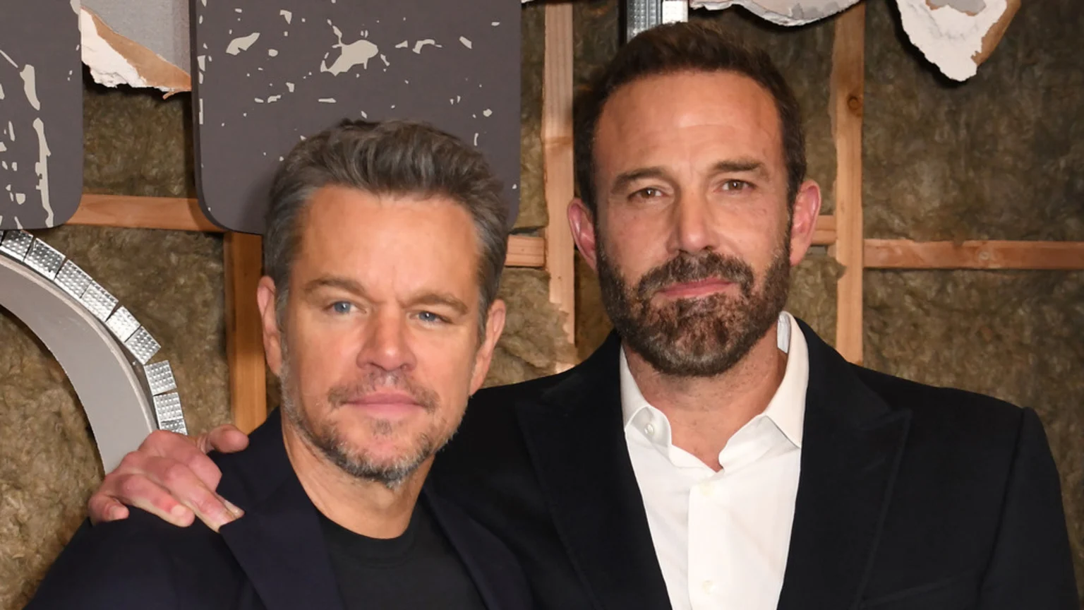 Actors Ben Affleck and Matt Damon pose for a photo in front of unfinished insulation, both bearded, with Damon's arm around Affleck's shoulder.