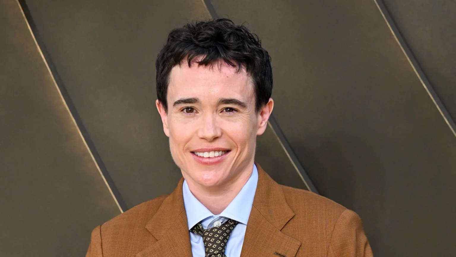 Elliot Page, with short dark curly hair, smiles at the camera while wearing a brown suit jacket, a light blue shirt, and a patterned tie.